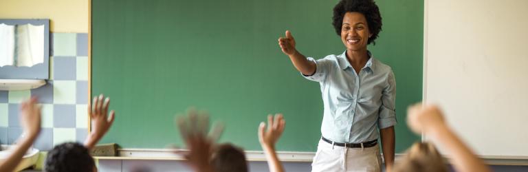 teacher standing at the front of the classroom pointing at students who have their arms raised