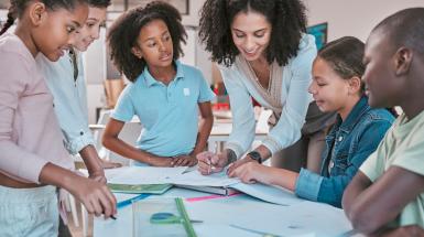 Female teacher in classroom with students, helping learner with homework and writing in book. Children's education at school, educator reading kids notebook and group learning together for assessment 